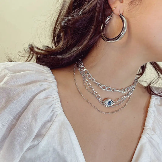 Close-up of a person wearing silver hoop earrings and layered necklaces with an eye pendant.