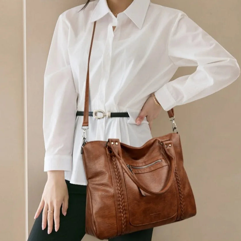 Person wearing a white shirt and holding a brown leather handbag against a beige background