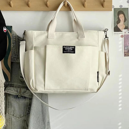 Beige tote bag with a strap hanging on a wooden hook against a white wall.
