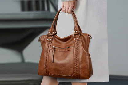 Brown leather handbag with braided detail held by a person, blurred background