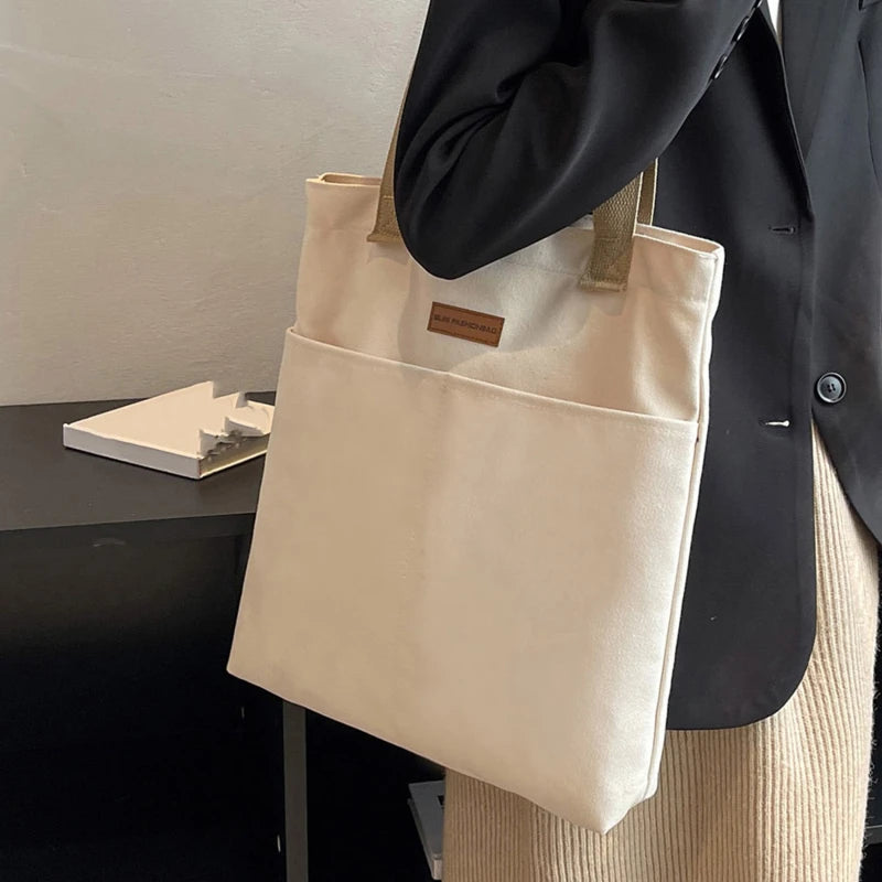 Beige tote bag held by a person wearing a black coat and beige pants, with a minimal background.