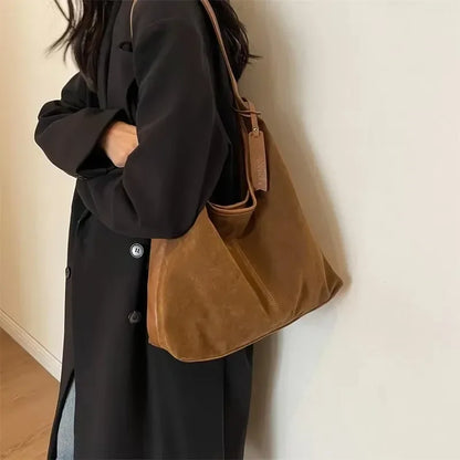 Person wearing a black coat with a brown leather bag over one shoulder against a plain background